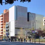 Edificio contemporáneo del Colegio de Economistas de Cataluña: fachada de cristal y paneles blancos, con su logotipo y nombre en el lateral derecho, situado junto a una avenida con peatones y arbolado